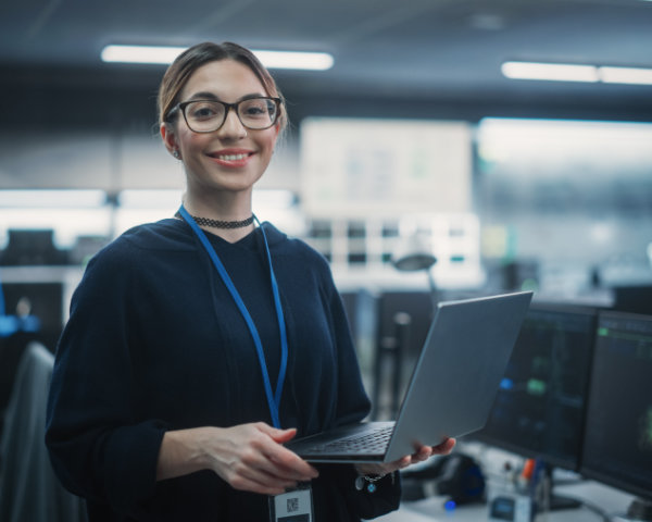 Non-binary IT professional holding a laptop while standing. They are wearing glasses, a blue sweater, and a lanyard with an access pass.