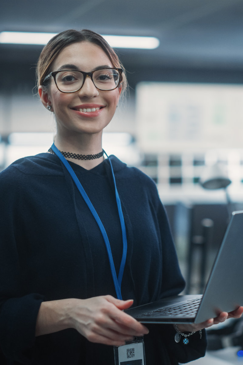 Non-binary IT professional holding a laptop while standing. They are wearing glasses, a blue sweater, and a lanyard with an access pass.