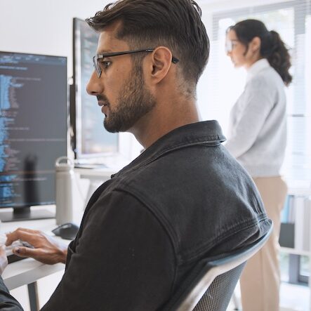 Close up side view of a man at a computer terminal focusing on solving a technical problem. A woman is in the background standing looking at her workstation.