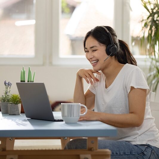Smiling woman sitting in a well-lit room at a table with a laptop in front of her. She is wearing a headset and participating in an online meeting.